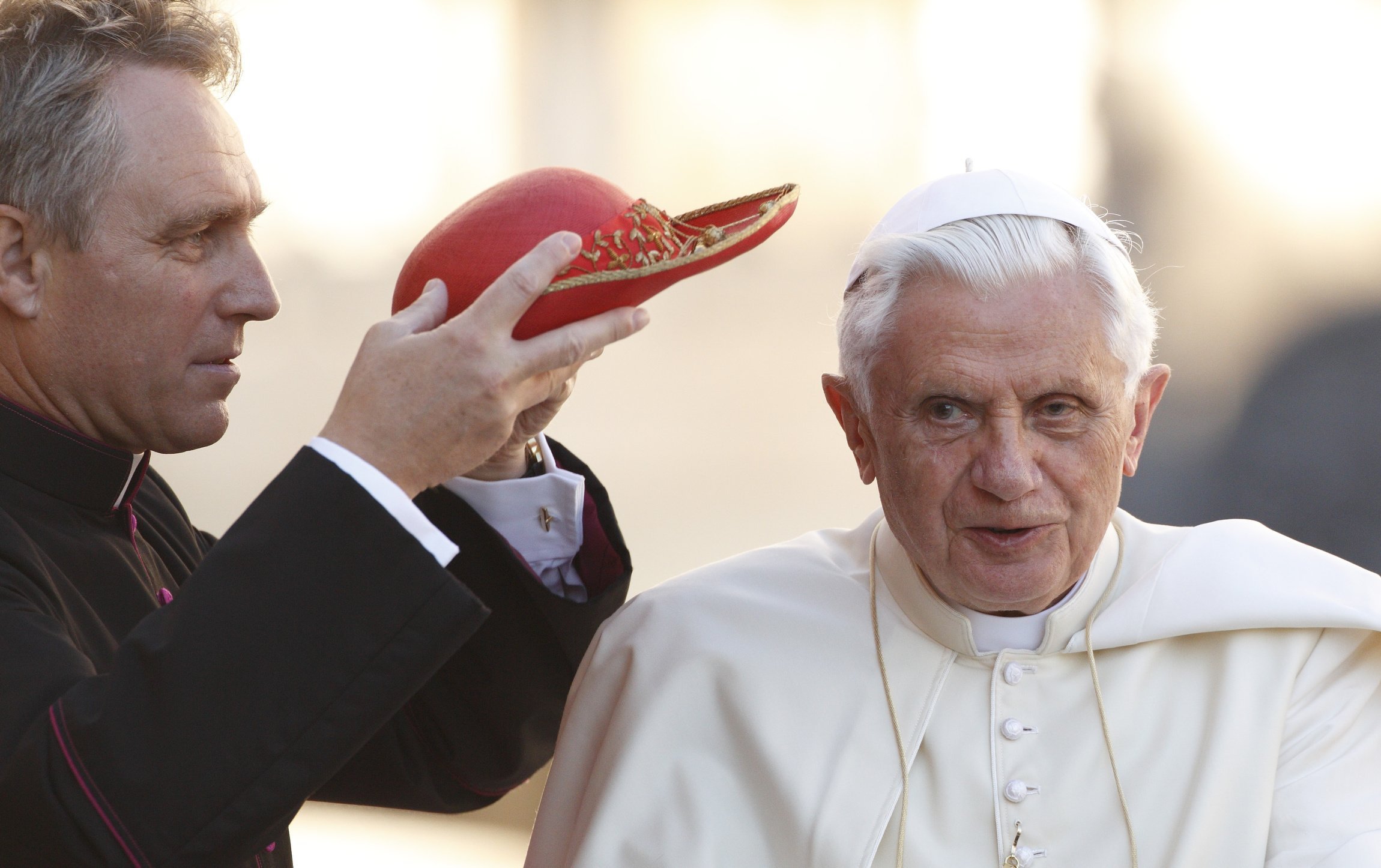 Then-Msgr. Georg Ganswein places a red hat on Pope Benedict XVI.