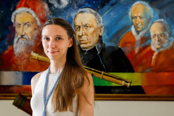 Student stands before a painting of popes and a telescope.